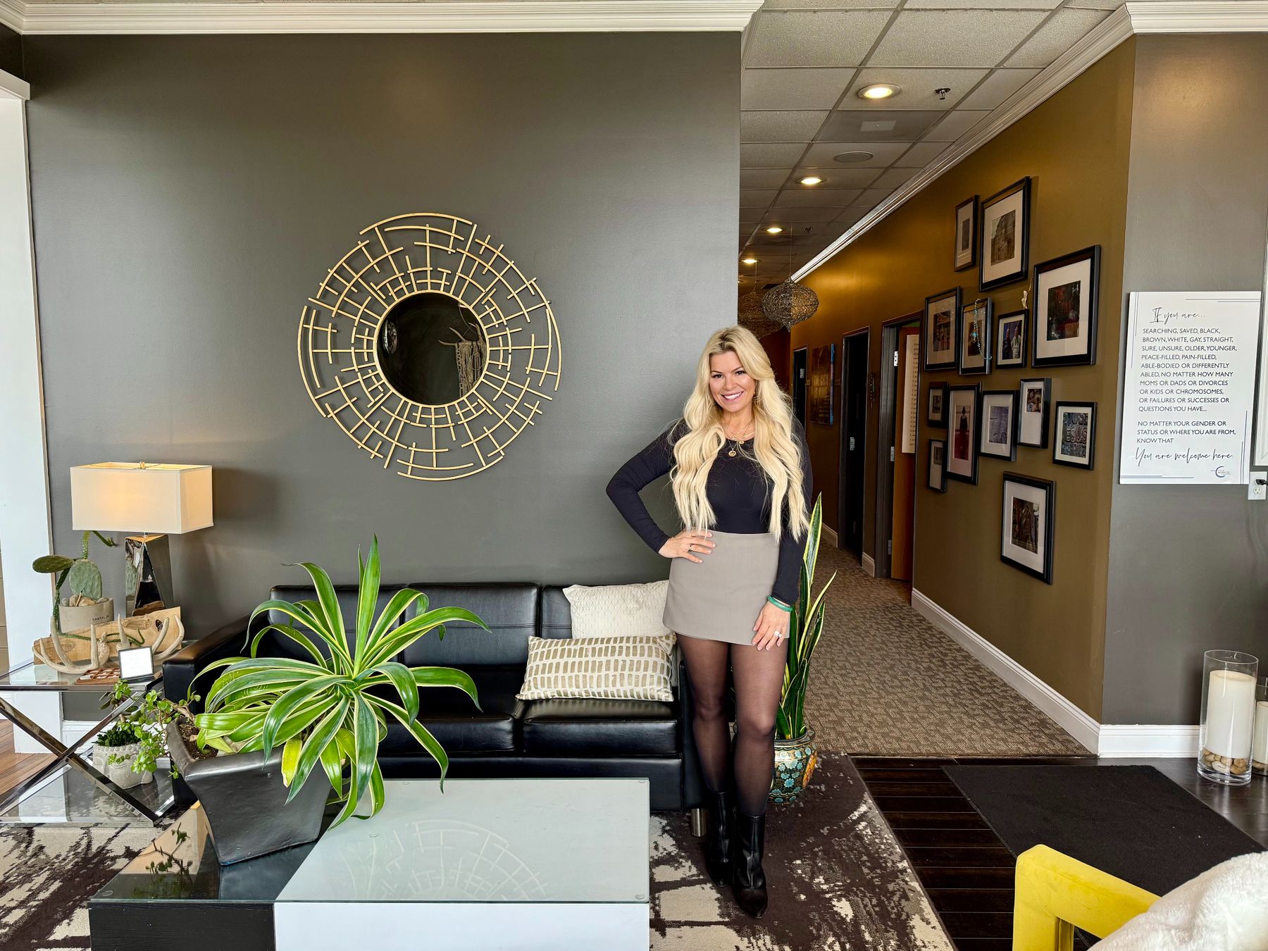 Dr. Christine Hanczaryk smiling in the HCNG lobby beside the brass sunburst mirror, with the black leather sofa, plants, and the gallery hallway behind her