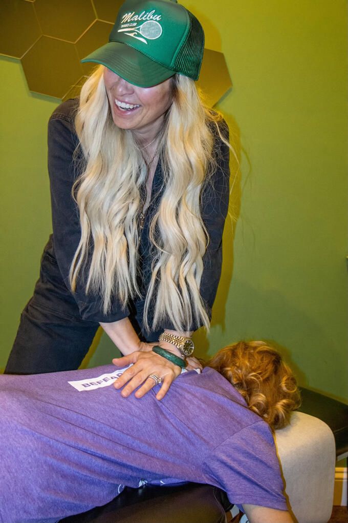 Dr. Christine, laughing under a green Malibu cap, adjusting a patient in front of the brass hex wall